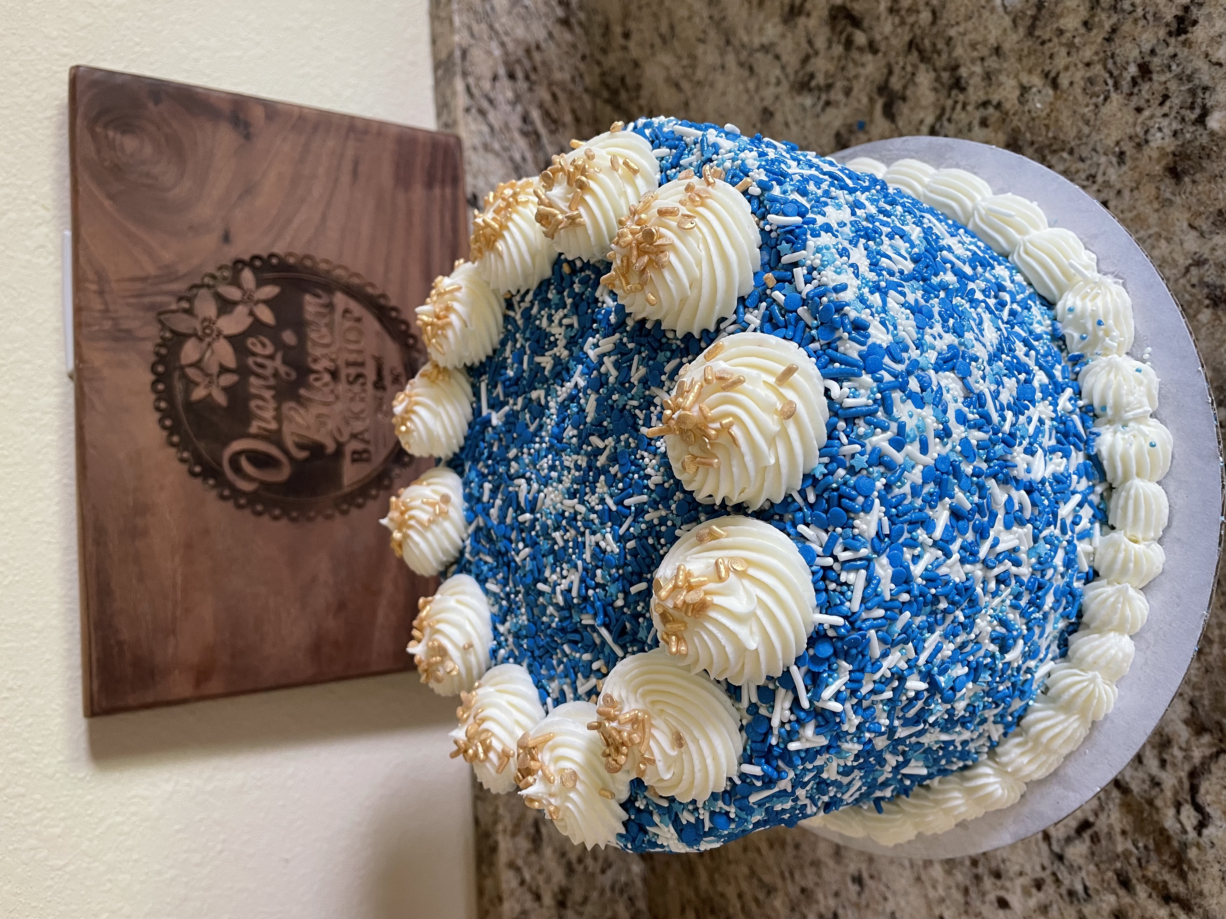 Blue celebration cake covered in blue sprinkles with white cream rosettes, Orange Blossom Bakeshop sign in background